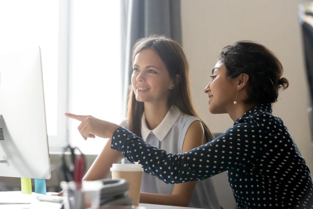 Smiling millennial diverse colleagues discussing online work together, happy caucasian female intern listening to indian mentor explaining computer task training employee, office mentoring concept (Smiling millennial diverse colleagues discussing onli