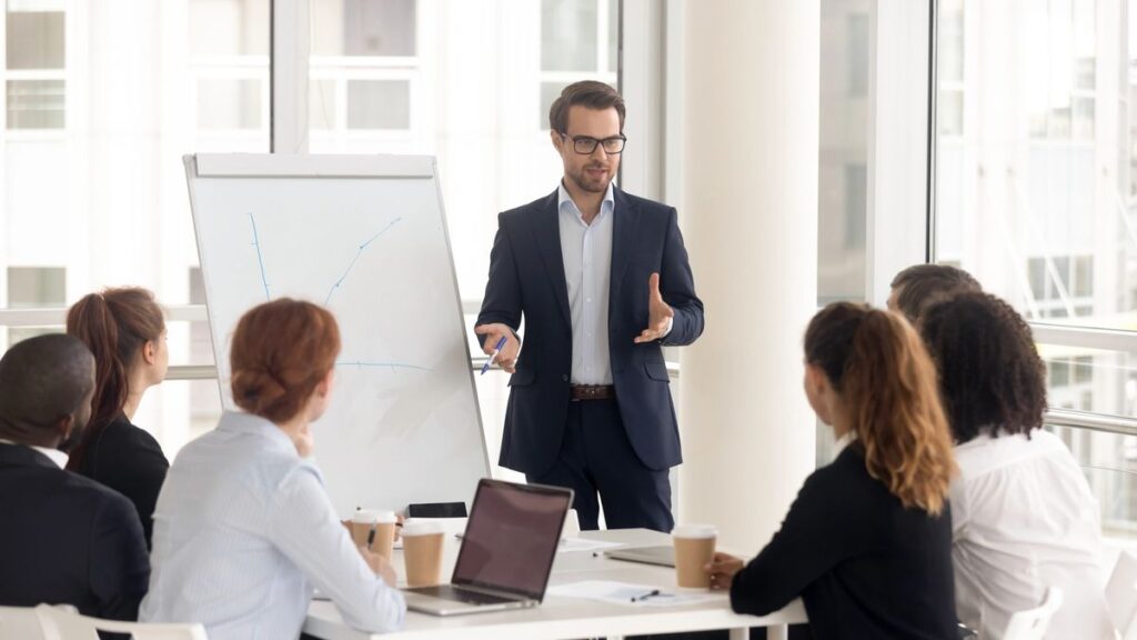 Male business coach speaker in suit give flipchart presentation, speaker presenter consulting training persuading employees client group, mentor leader explain graph strategy at team meeting workshop (Male business coach speaker in suit give flipchart