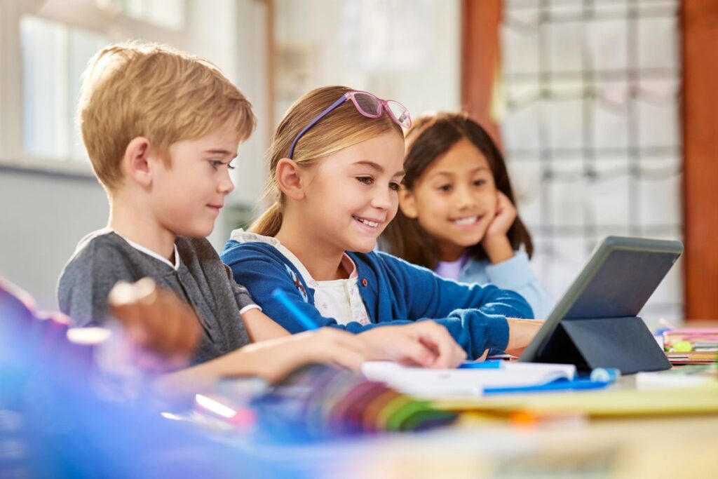 Happy multiethnic elementary classmates working on digital tablet in classroom during lesson. Three elementary school pupils using digital tablet at desk during technology class. Schoolchildren in class using technology for studying together in a inte